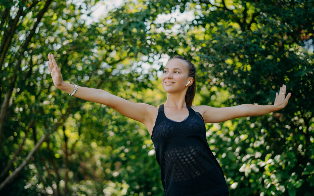 Training outdoors concept. Pleased sporty European woman feels freedom motivates you for sport stretches arms sideways leads active lifestyle wears black t shirt poses against green trees outside.