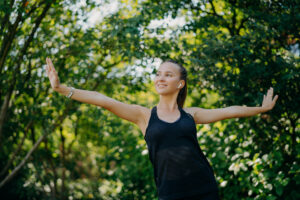 Training outdoors concept. Pleased sporty European woman feels freedom motivates you for sport stretches arms sideways leads active lifestyle wears black t shirt poses against green trees outside.