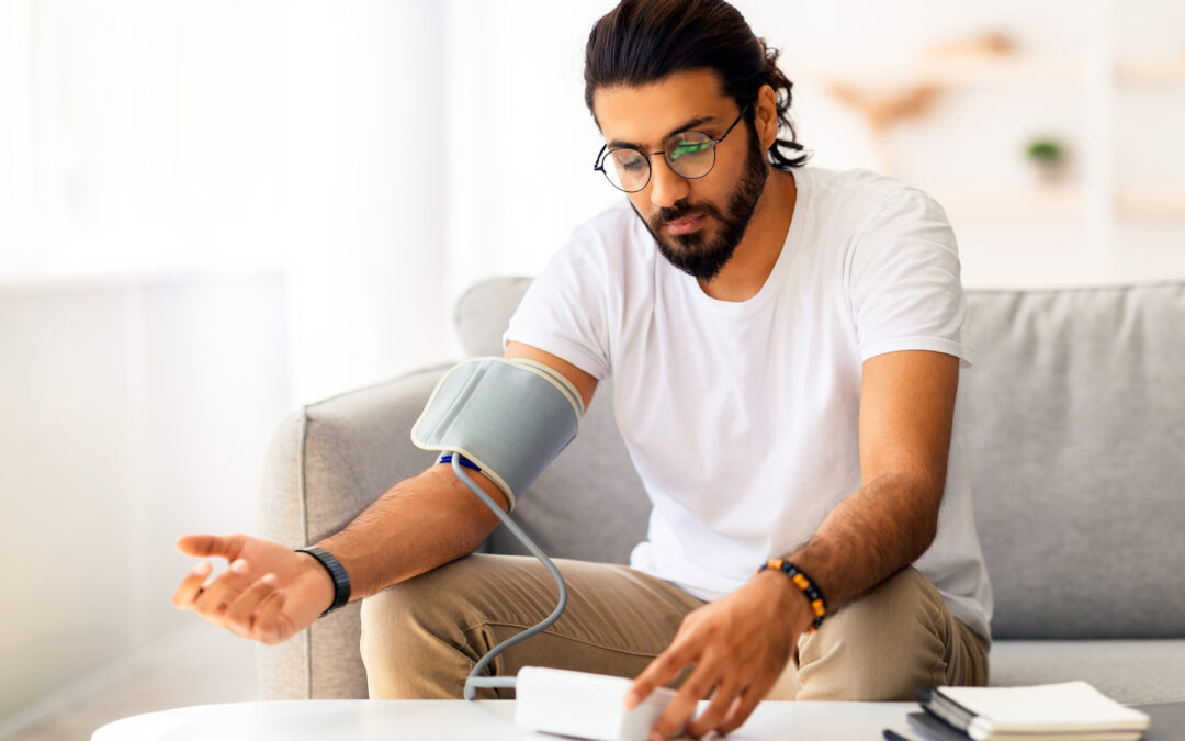 Young man sitting on couch, checking blood pressure and taking notes at home. Indian guy in white t-shirt using modern tonometer, experiencing hypertension caused by stress, copy space